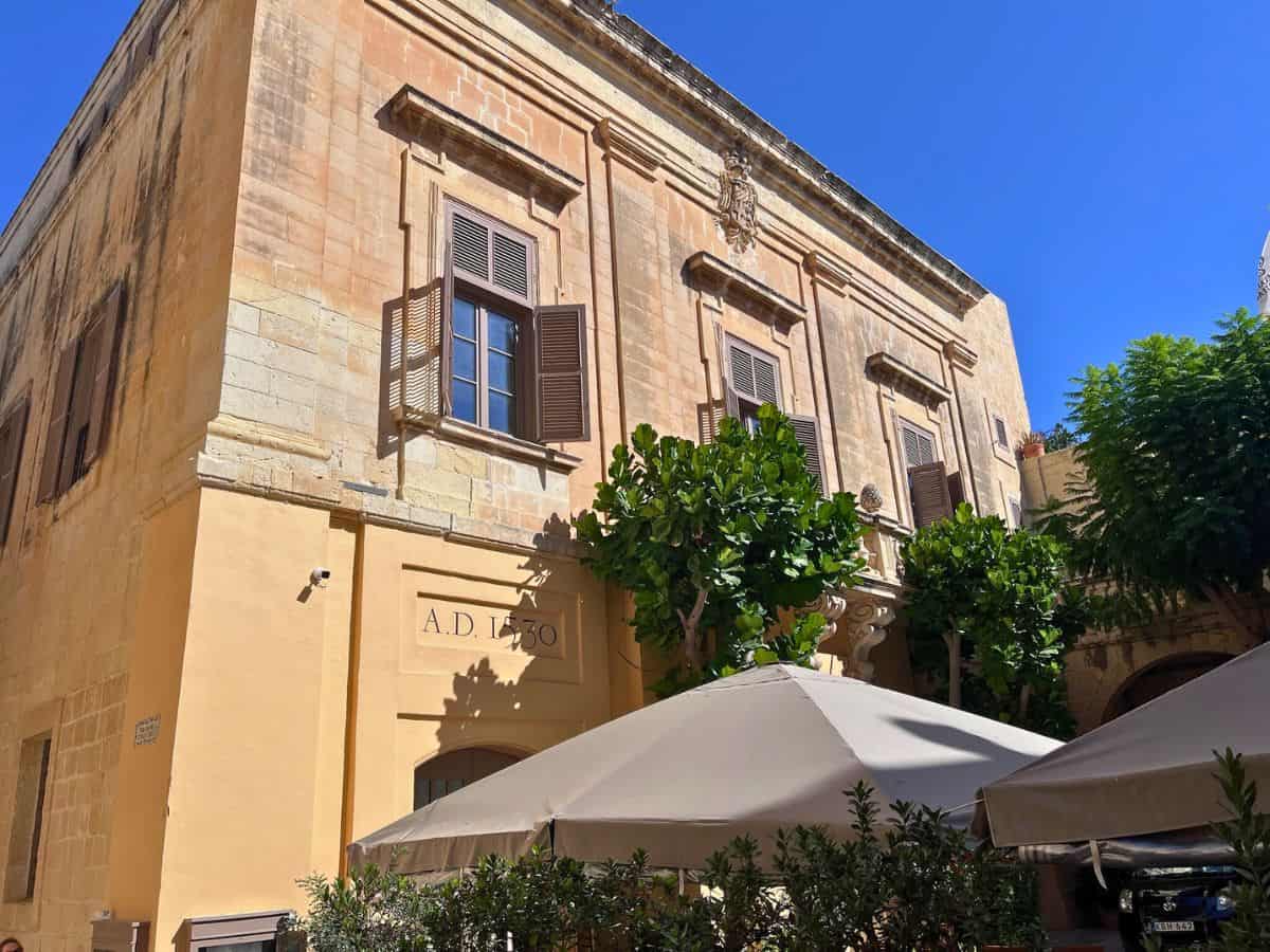 A honey-colored limestone building (typical of Malta) with cafe umbrellas